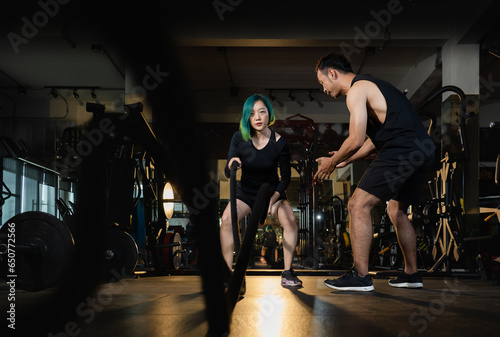 asian athletic woman exercise crossfit battle rope with young personal trainer cheer up at modern gym, active fitness people workout weight training strength body and mental health in dark sport club