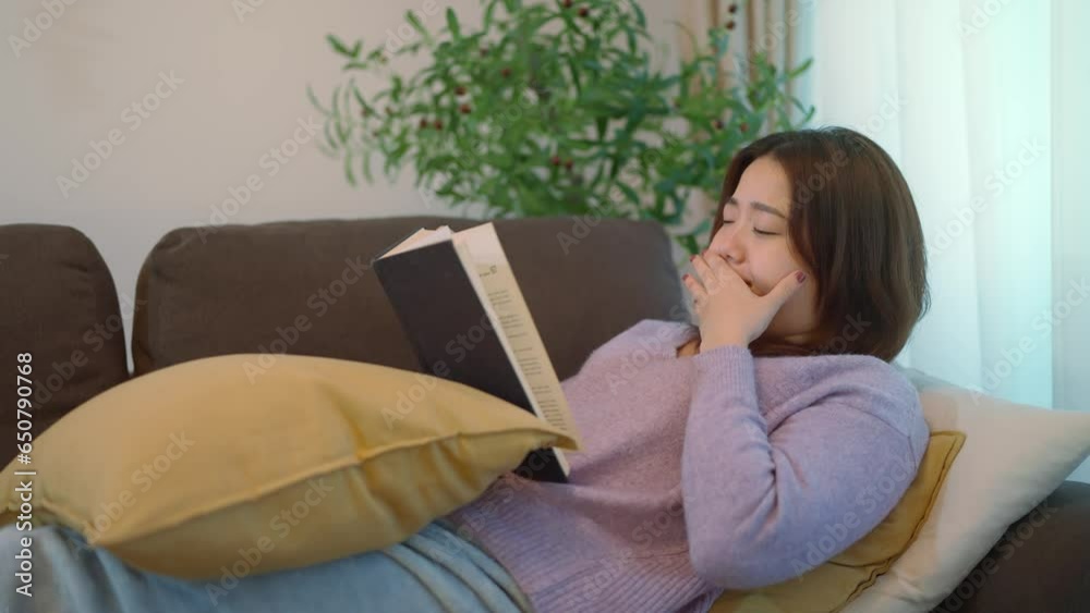 Young asian woman reading a book and sleepy while laying on couch in living room. Sleepy women reading a book while relaxing on couch on weekend. Lifestyle concept Young asian woman reading a book and sleepy while laying on couch in living room. Sleepy women reading a book while relaxing on couch on weekend. Lifestyle concept