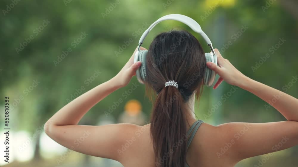 Happy asian woman adjusting wireless headphones before starting jogging and listening to music on running route surrounded by nature. Slow motion.