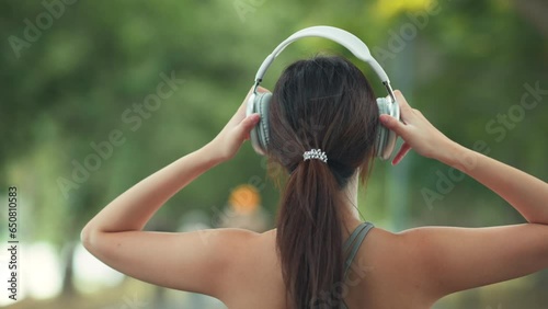Happy asian woman adjusting wireless headphones before starting jogging and listening to music on running route surrounded by nature. Slow motion. photo