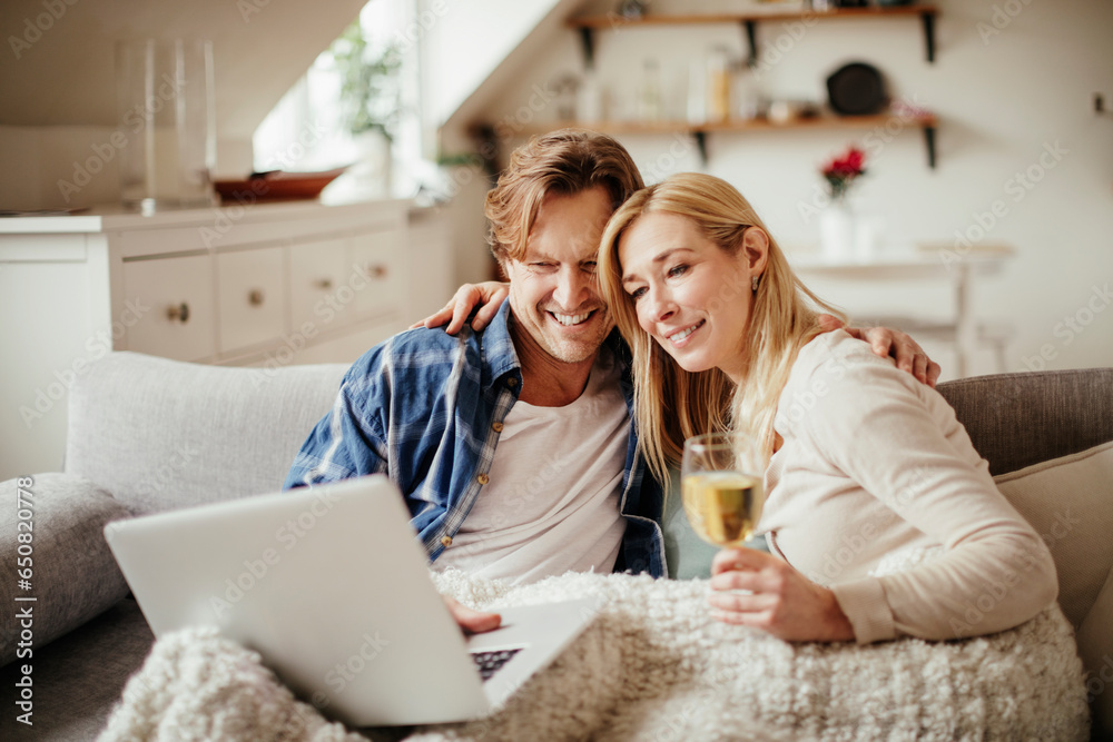 Middle aged Caucasian couple having wine while using a laptop under a blanket in the living room Middle aged Caucasian couple having wine while using a laptop under a blanket in the living room