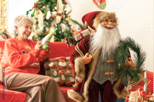 La hermosa abuela celebra la Navidad en casa, sosteniendo un adorno navideño en mano, mientras la figura decorativa de Papá Noel sonríe en primer plano, iluminando el espíritu festivo del hogar photo