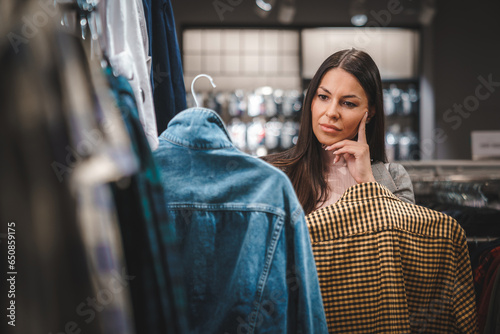 Attractive young woman shopping and feeling hesitant while trying to decide between two different pieces of clothes in a retail store. Pensive undecided woman making choices in a shopping mall.