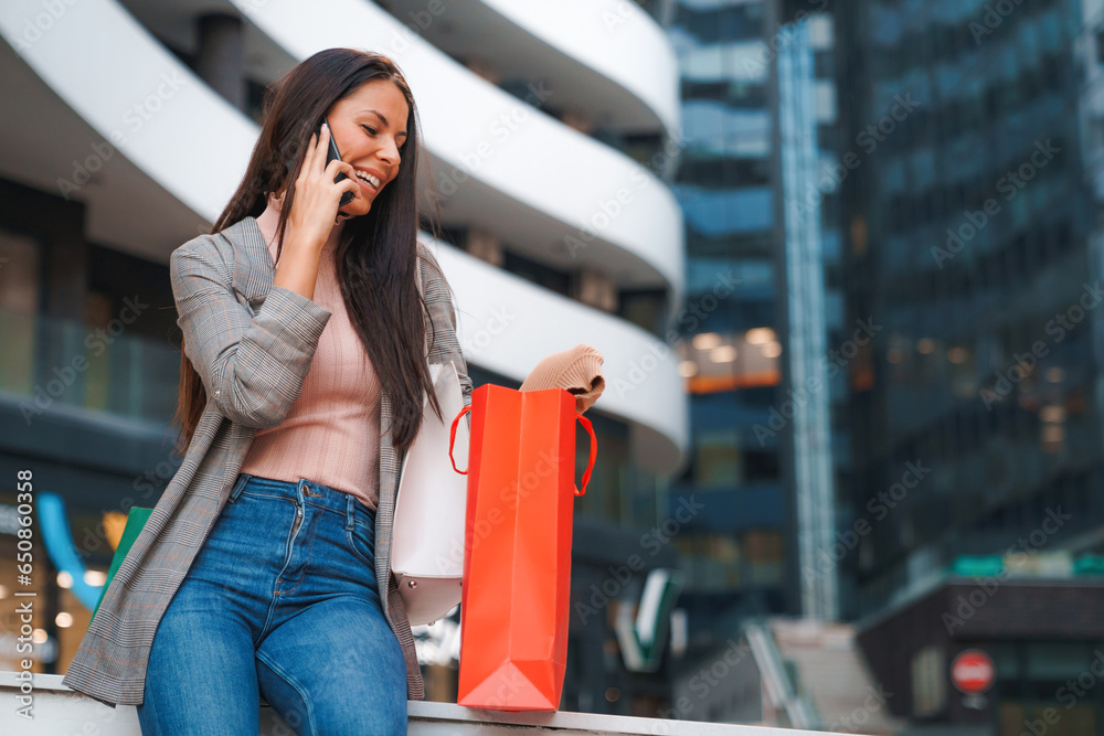 Fototapeta premium Beautiful young business woman sitting in front of modern shopping mall, having a phone call and looking at purchased clothes in shopping bag.