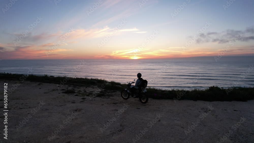 Aerial video of motorcyclist on a mountain peak looking at the sea during sunset Aerial video of motorcyclist on a mountain peak looking at the sea during sunset