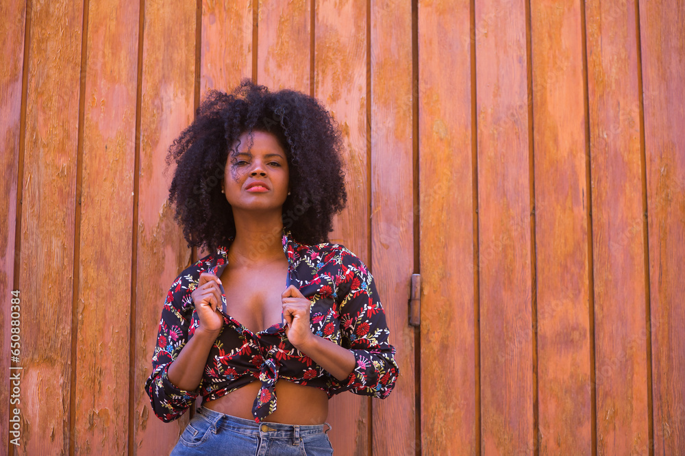 Fototapeta premium Young woman, beautiful and black with afro hair, with a floral shirt leaning on a wooden board door, in a sensual, provocative and flirtatious attitude. Concept beauty, sensuality, provoke, flirt.