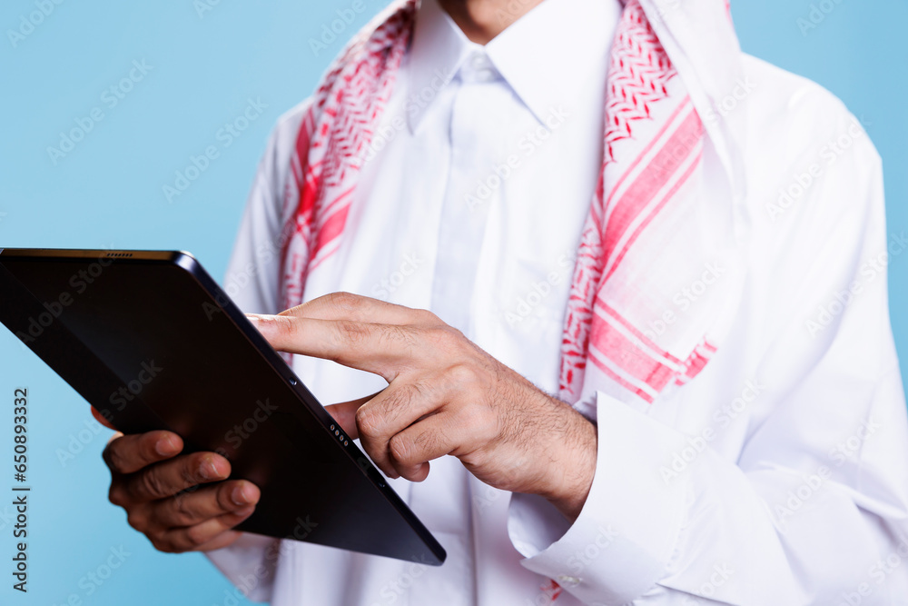Man dressed in traditional muslim clothes holding digital tablet in hands and tapping on screen closeup. Person wearing thobe using portable gadget and browsing internet page Man dressed in traditional muslim clothes holding digital tablet in hands and tapping on screen closeup. Person wearing thobe using portable gadget and browsing internet page