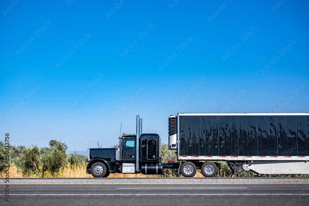 Brilliant black big rig classic semi truck tractor transporting cargo in covered reefer semi trailer driving on the flat horizontal highway road