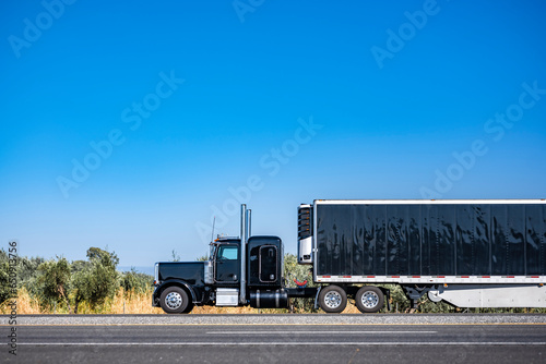 Brilliant black big rig classic semi truck tractor transporting cargo in covered reefer semi trailer driving on the flat horizontal highway road