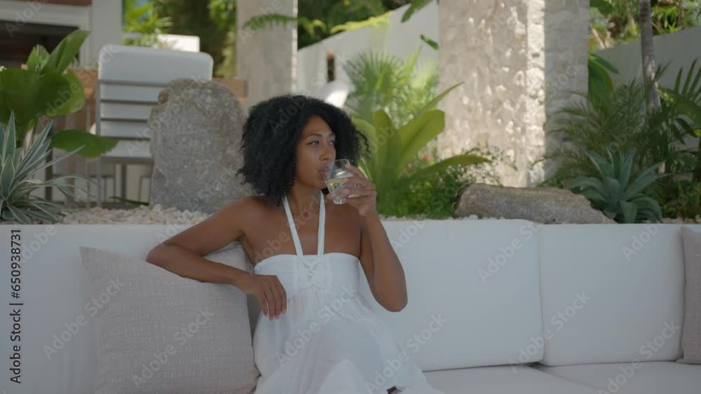 Beautiful black woman with short hair dressed in white sundress sitting on white sofa and drinking water enjoying her time, exotic holidays concept
