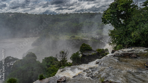 The raging stream of the river collapses from the ledge into the abyss. Spray and fog. Lush tropical vegetation all around. Clouds in the sky. Iguazu Falls. Argentina.