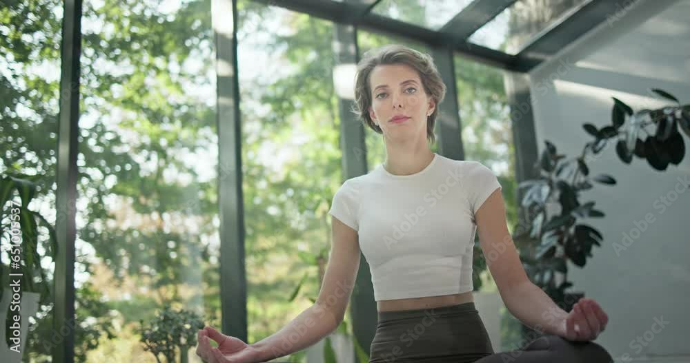young caucasian fitness woman sitting in lotus position on a yoga mat in a modern studio with plants and large forest or garden overlooking windows young caucasian fitness woman sitting in lotus position on a yoga mat in a modern studio with plants and large forest or garden overlooking windows