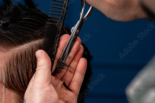 Focused Caucasian male hairdresser cuts a Caucasian guy s hair in a beauty salon. hairdresser at work  blue background  haircut with a typewriter and scissors. 