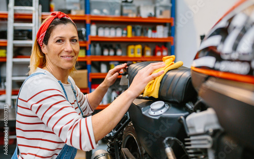 Portrait of smiling female mechanic with freckles and hair bandana looking at camera while cleaning motorcycle seat with a microfiber cloth on factory