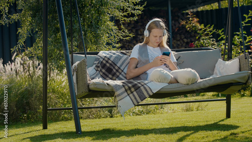 A teenage girl is resting on a garden swing, using a smartphone.