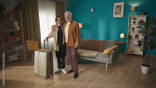 Shot of a teenager, young man with his grandfather, granddad having a photoshoot together, hugging and smiling at the camera. The boy holds a suitcase. photo