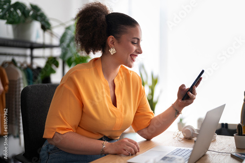 Portrait of content female fashion designer smiling and looking at mobile phone while working at table with laptop in atelier photo