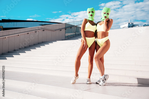 Two beautiful sexy women in green underwear. Models wearing bandit balaclava mask. Hot seductive female in nice lingerie posing in the street at sunny day, blue sky. Crime and violence. Perfect body