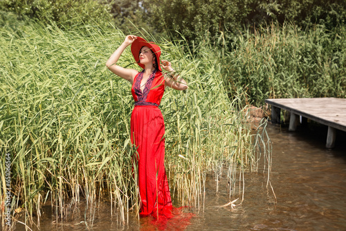 A beautiful elegant lady in a red hat and a long red dress walks along the water along the shore with reeds. emotional portrait of a girl in the water