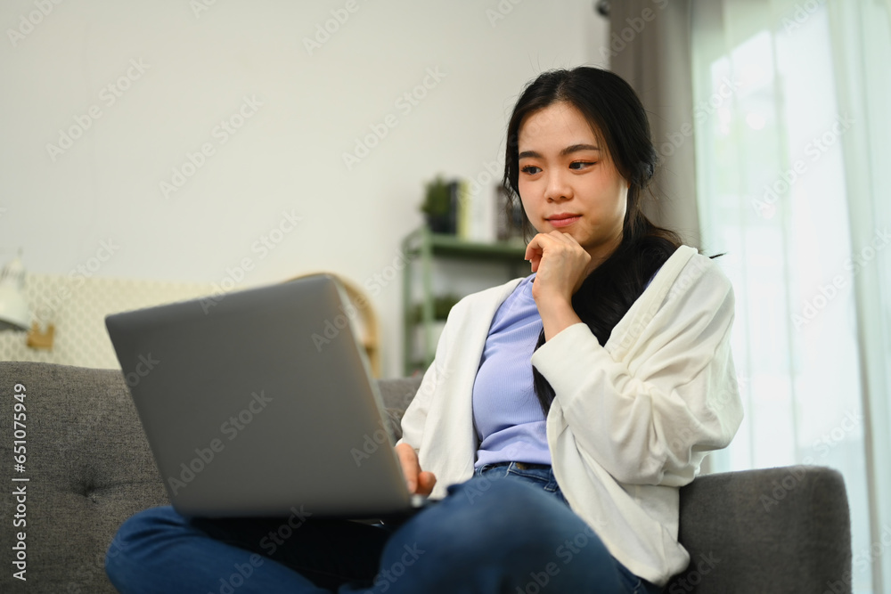 Carefree young woman sitting on couch at home and using laptop, browsing internet or shopping online Carefree young woman sitting on couch at home and using laptop, browsing internet or shopping online