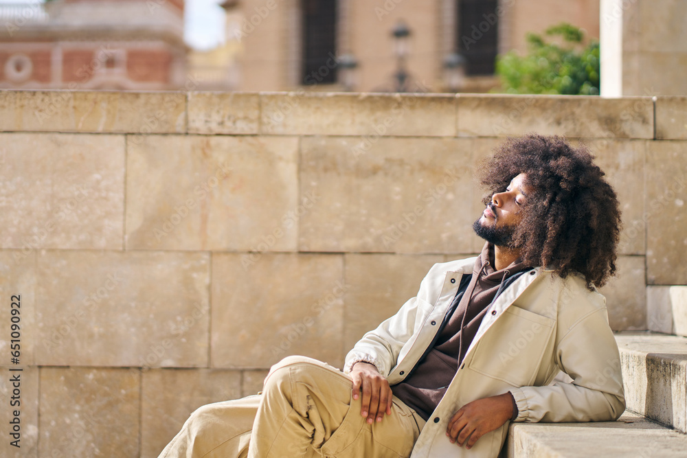 afro man sitting in a square enjoying the sun on his face concept of wellbeing and inner peace