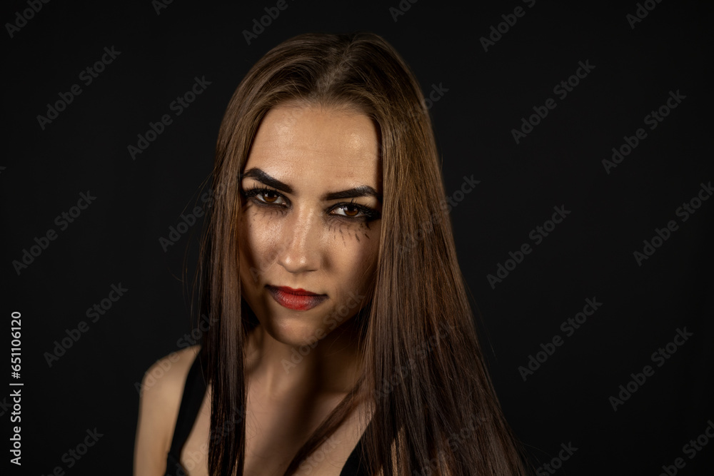 Portrait of a beautiful witch on a black background. A woman with long black hair and black makeup looks seriously into the camera. Woman witch sternly looks into the camera on a black background.