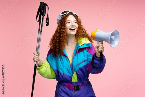 Happy curly haired woman, skier wearing stylish winter overalls, ski goggles holding megaphone isolated on pink background. Shopping, sale announcement concept