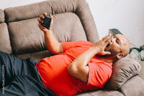 Lazy man yawns while browsing the internet or watching movies on his phone, lying lazily on the couch.