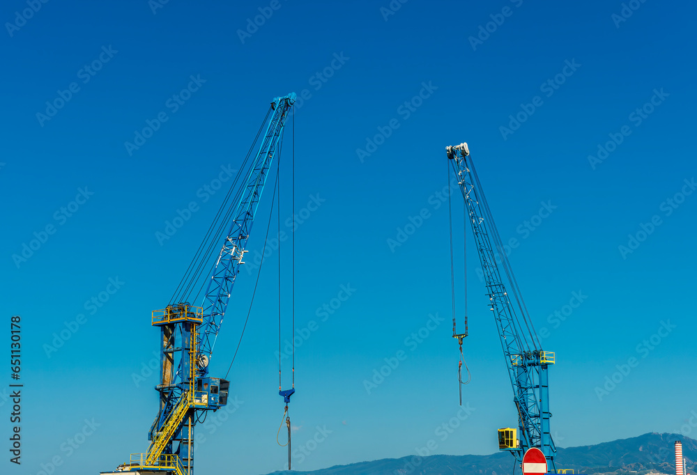 Two construction tower cranes at a construction site against a blue sky with clouds. Two construction tower cranes at a construction site against a blue sky with clouds.