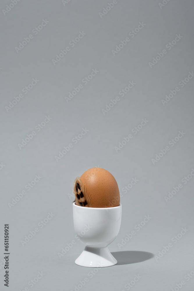 Brown egg in white eggcup with brown feather. Gray background, space for text. Minimal Easter card. Brown egg in white eggcup with brown feather. Gray background, space for text. Minimal Easter card.