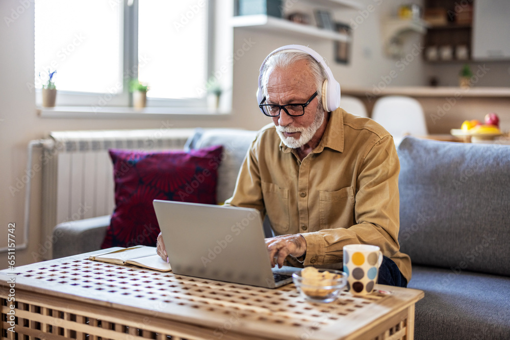 Portrait of senior man learning online with headphones and laptop at home, technology and leisure concept. Older mature man using wireless laptop apps browsing internet sit on sofa. - obrazy, fototapety, plakaty 