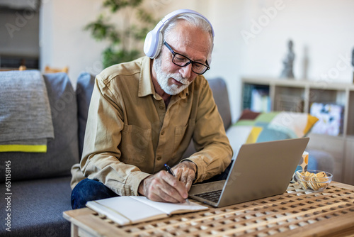 Portrait of senior man learning online with headphones and laptop at home, technology and leisure concept. Older mature man using wireless laptop apps browsing internet sit on sofa.