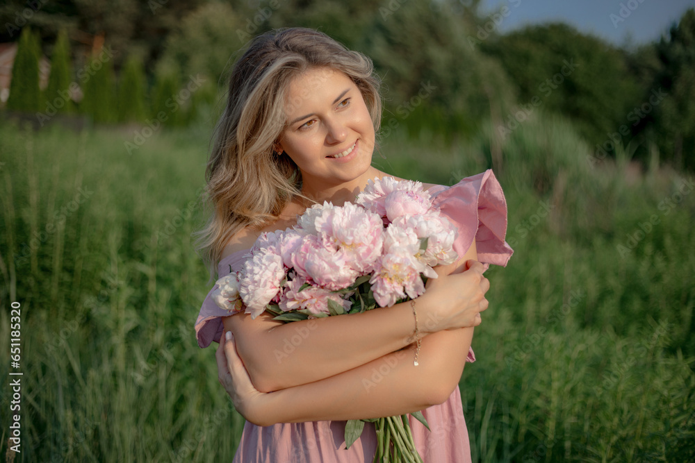 Fototapeta premium Charming natural blonde with loose hair in pink romantic dress with bouquet of peonies in nature. 