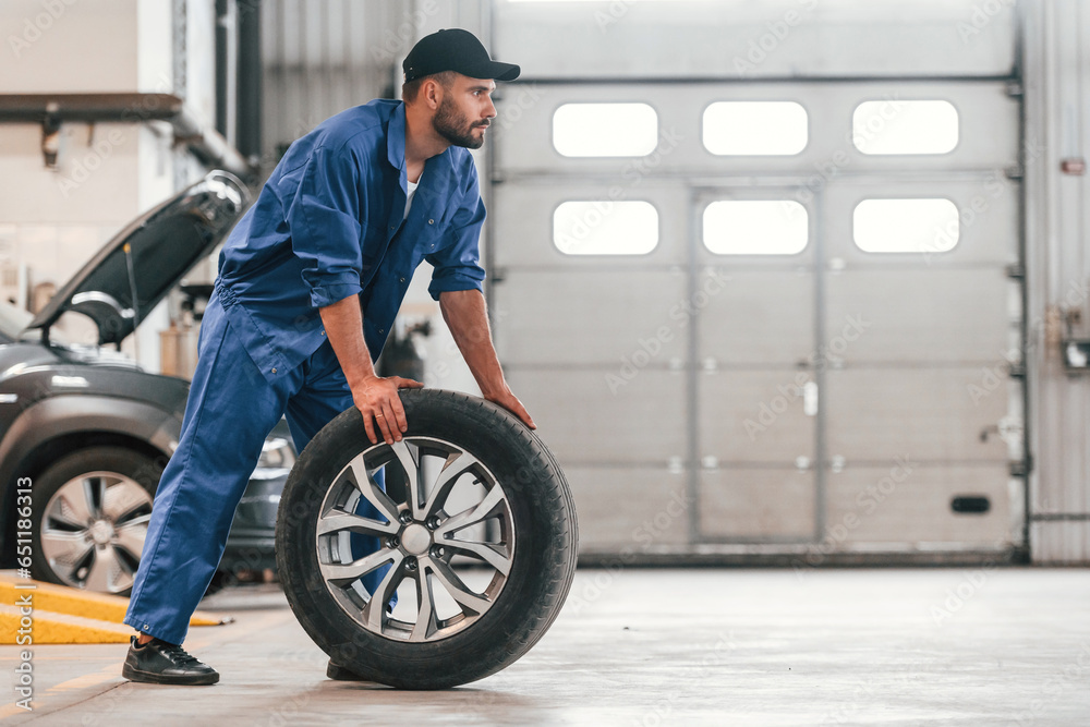 Side view. Moving the new tire. Auto mechanic working in garage. Repair service Side view. Moving the new tire. Auto mechanic working in garage. Repair service