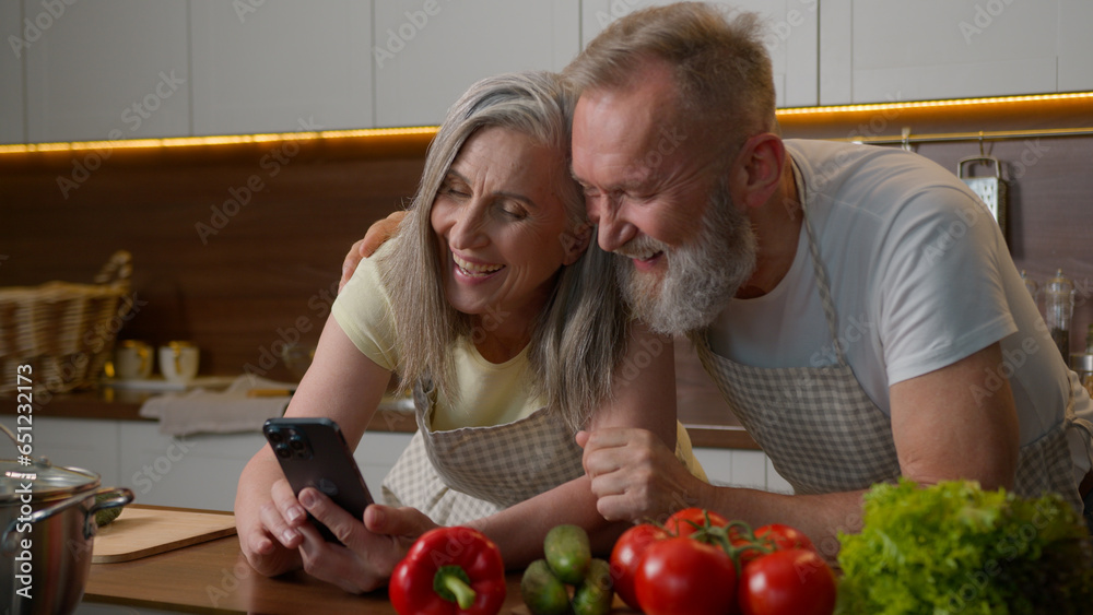 Laughing retired old gray haired Caucasian family love couple using mobile phone online order delivery food vegetables on kitchen table happy senior woman and man talking shopping cooking healthy meal Laughing retired old gray haired Caucasian family love couple using mobile phone online order delivery food vegetables on kitchen table happy senior woman and man talking shopping cooking healthy meal