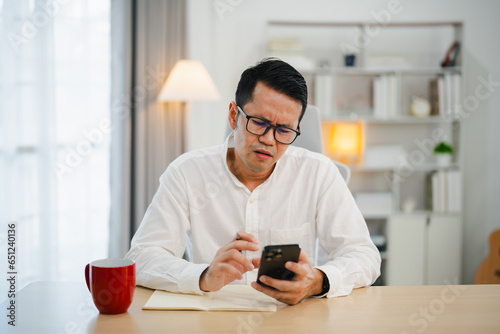 Depression concept. Asian man wearning glasses sad and checking mobile phone sitting on the table in the living room at home, Asian man serious and worry while using mobile phone. photo