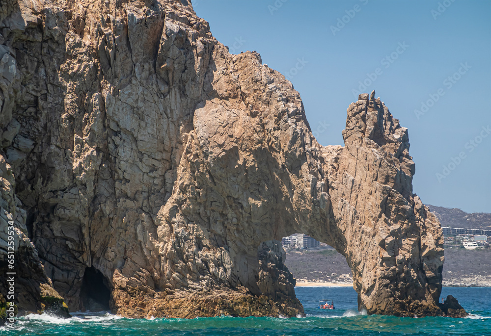 Mexico, Cabo San Lucas - July 16, 2023: El Arco from ocean side as part of brown-beige rocky cliffs, with small boat captured in its arch. Cityscape on horizon