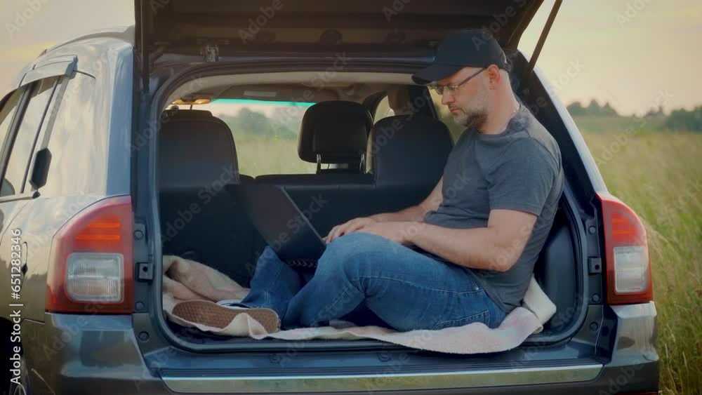 Man working remotely on laptop in nature sitting in car trunk while traveling. Busy worker freelancer entrepreneur notebook online working outdoors in SUV Man working remotely on laptop in nature sitting in car trunk while traveling. Busy worker freelancer entrepreneur notebook online working outdoors in SUV