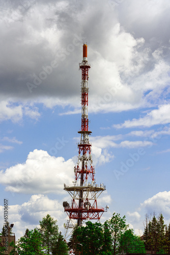 A tall red and white telecommunications tower against a blue sky with white clouds. The tower has a lattice structure with antennas and dishes. At the top there is a lighthouse, surrounded by trees