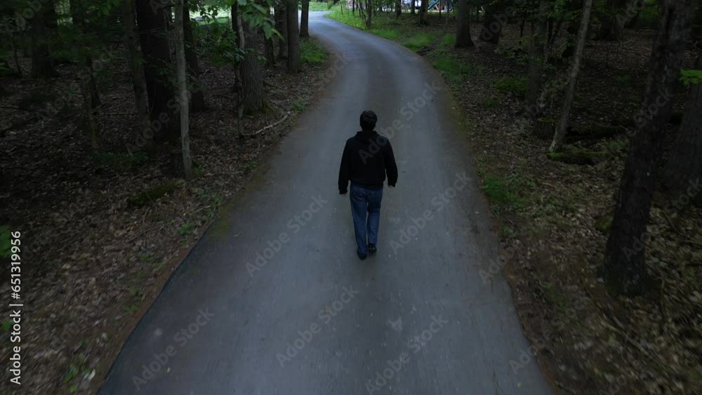 aerial footage of man walking away from behind on small paved road, driveway surrounded by trees in rural suburban area (4k drone shot from above) following flying