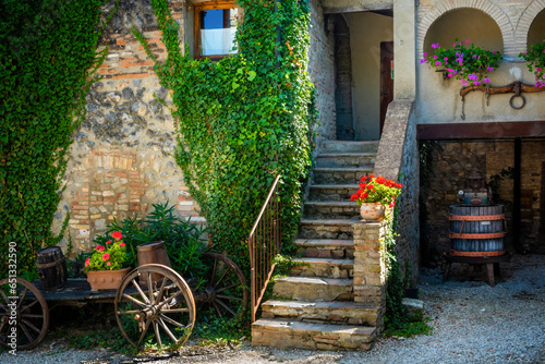A peaceful Tuscany farmyard with stairs leading up into the house  a wine crushing barrel  and a cart. The summer light falls on greenery and red flowers.