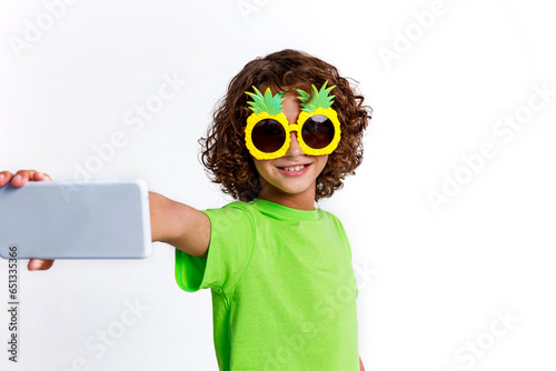 Positive curly kid boy take selfie wear green t-shirt and funny sunglasses on white background