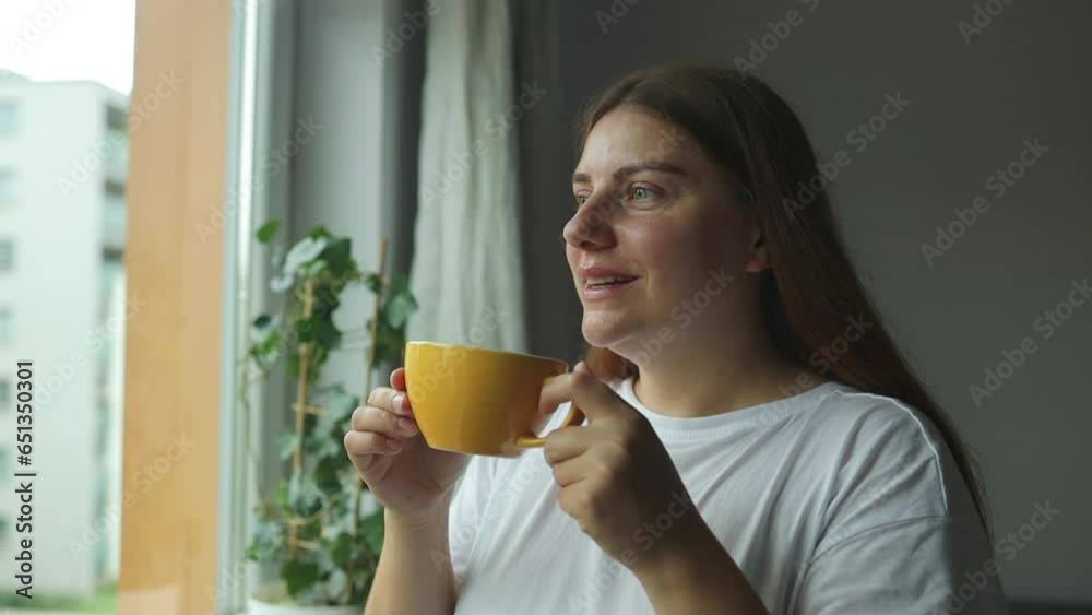 Beautiful relaxed woman with cup of tea or coffee and looking through window while sitting at windowsill at home. High quality FullHD footage Beautiful relaxed woman with cup of tea or coffee and looking through window while sitting at windowsill at home. High quality FullHD footage
