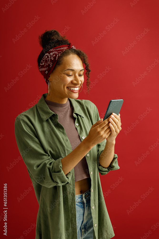 Pretty woman is using mobile phone with smile while standing on red studio background Pretty woman is using mobile phone with smile while standing on red studio background