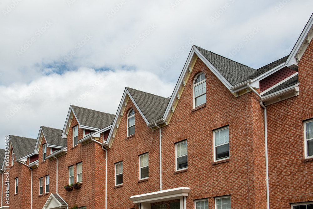 A row of brown brick townhouses with peaked gable roofs, and double hung windows with white trim. The shingles are black asphalt. The adjoined facade row houses are multi-story residential buildings. 