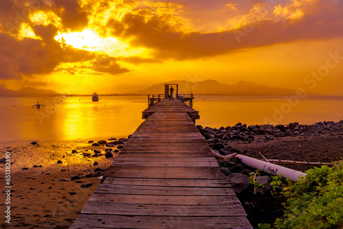 A wooden pier juts out into a calm body of water, bathed in the golden light of sunrise. © Neilstha Firman