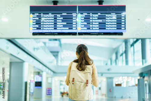 Happy beautiful Asian female backpacker tourist standing and looking at flight departure and arrival information at the airport terminal.