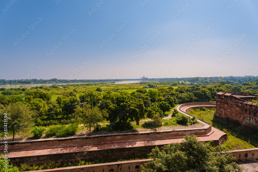  beautiful white marble Taj Mahal is seen from the Red Fort . In the foreground are green fields, a river,  blue sky. India, Agra.