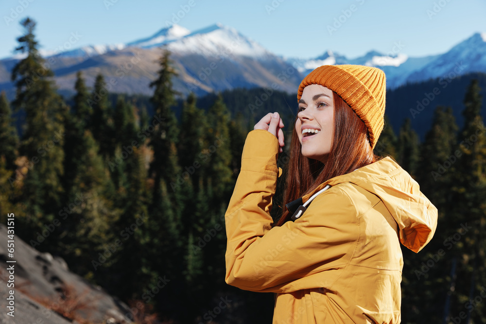 Woman smiling with teeth looking at the mountains in a yellow raincoat with red hair on a hike standing in front of the mountains in a yellow winter sunset cap