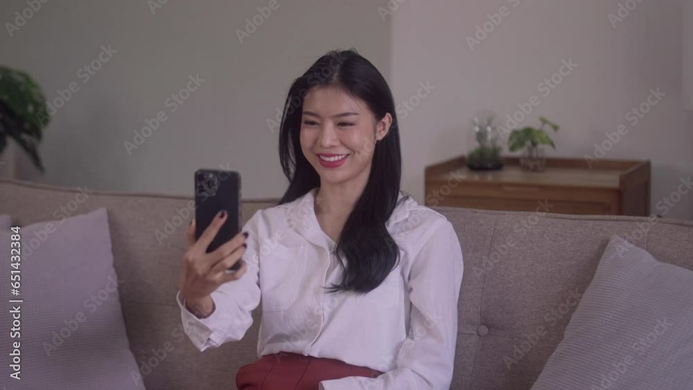 smiling young attractive Asian woman using smartphone waving hand for streaming stories online in social networks in conversation on couch at home. smiling young attractive Asian woman using smartphone waving hand for streaming stories online in social networks in conversation on couch at home.